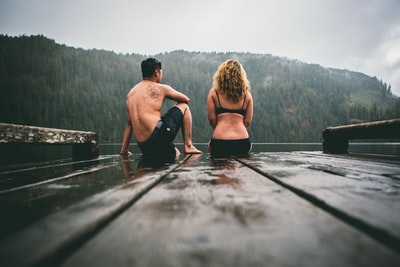 在灰色的天空下，一对男女坐在棕色的木船码头上，面对着水体
